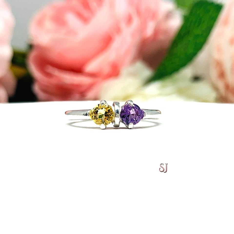 A sterling silver mother's ring featuring a 4mm round golden citrine and 4mm round amethyst separated by a rounded bar. Shown displayed on a white surface with a background of pastel pink and white flowers.
