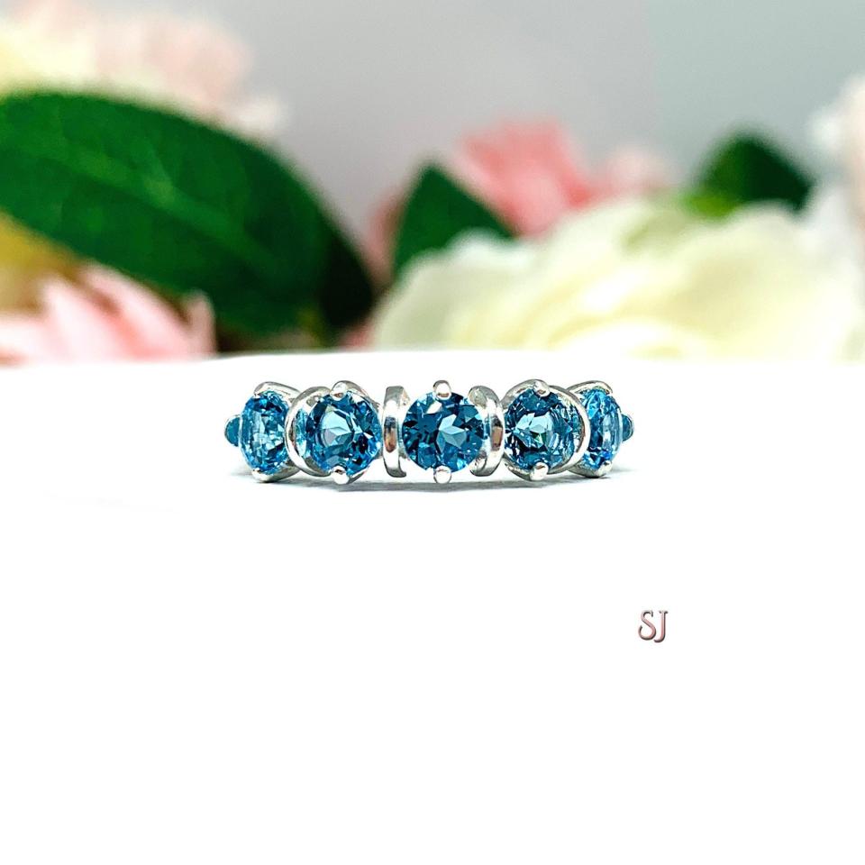 A sterling silver ring featuring five 4mm round swiss blue topaz gemstones in a row. The ring has rounded bars separating each stone. Displayed on a white surface with pastel floral background.