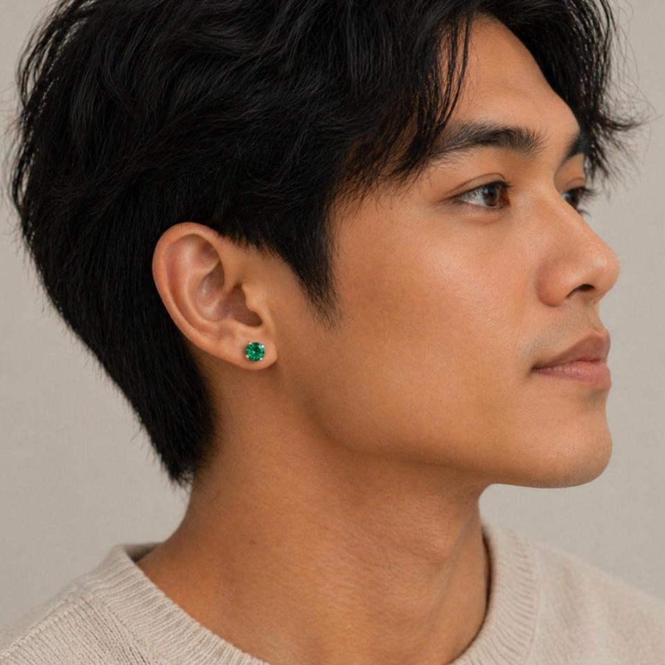 Man wearing sterling silver stud earrings showcasing vibrant emerald green round moissanite stones in prong settings. Model image for size and styling reference — not a photo of the actual earrings.