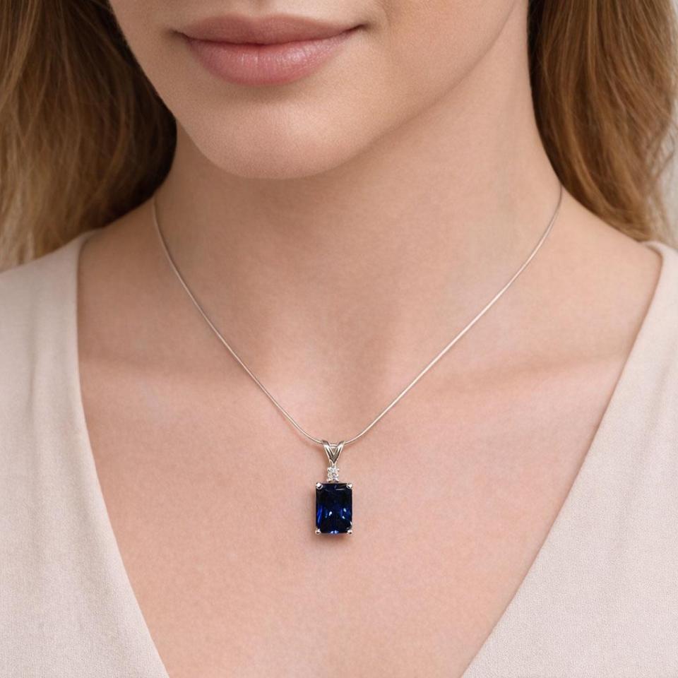 Close‑up of a woman wearing a sterling silver necklace with a 9x7mm radiant‑cut lab‑created blue sapphire pendant accented by a 2.5mm round moissanite. The pendant rests just below her collarbone on a thin silver chain. She wears a neutral beige top, and the lighting softly emphasizes the sapphire’s deep blue color and sparkle. Model image for size and styling reference — not a photo of the actual necklace.