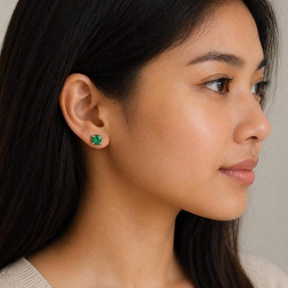 Woman wearing sterling silver stud earrings showcasing vibrant emerald green round moissanite stones in prong settings. Model image for size and styling reference — not a photo of the actual earrings.
