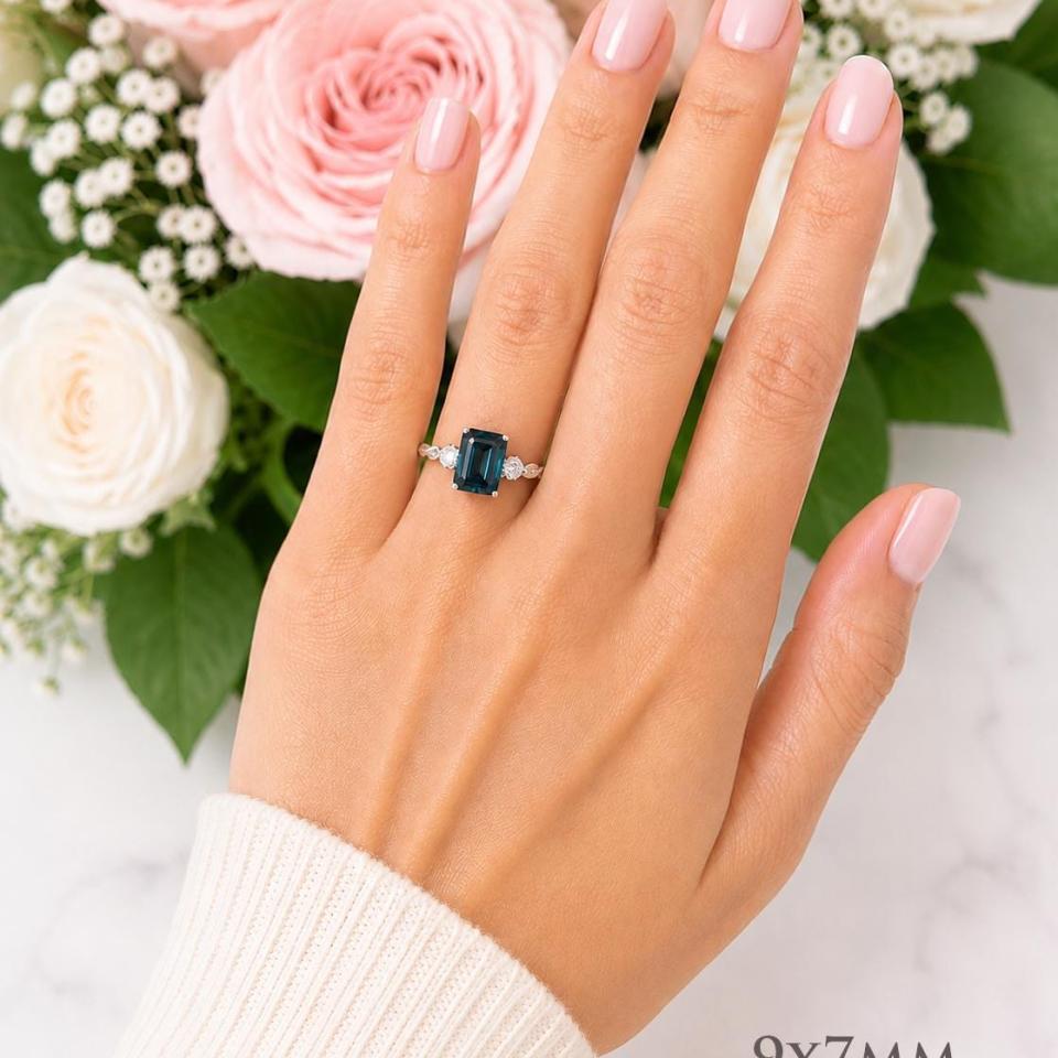 A woman's hand wearing a sterling silver ring featuring a 9x7mm emerald cut London blue topaz flanked by a round moissanite on each side. Model image for size and styling reference — not a photo of the actual ring.