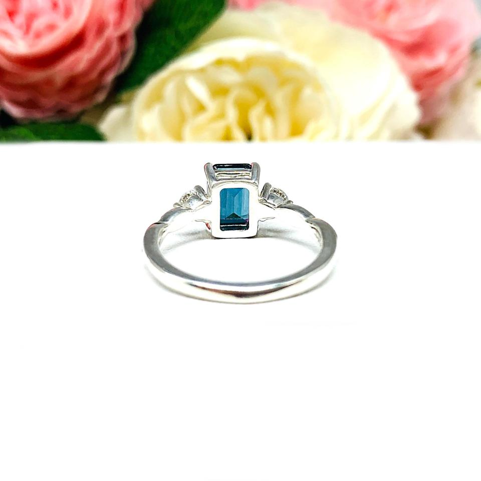 Close-up of an 8x6mm emerald-cut London Blue Topaz ring with moissanite side stones, set in a sterling silver twist band with milgrain detailing; soft floral background enhances the elegance.
