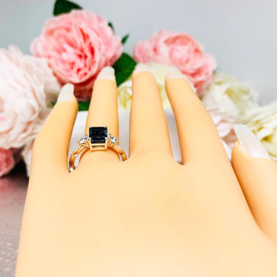 Mannequin hand with long white shimmery nails, displayed against a soft white-gray background with pink and white floral accents. The hand is wearing an 8x6mm emerald-cut London Blue Topaz ring with moissanite side stones, set in a sterling silver twist band with milgrain detailing.