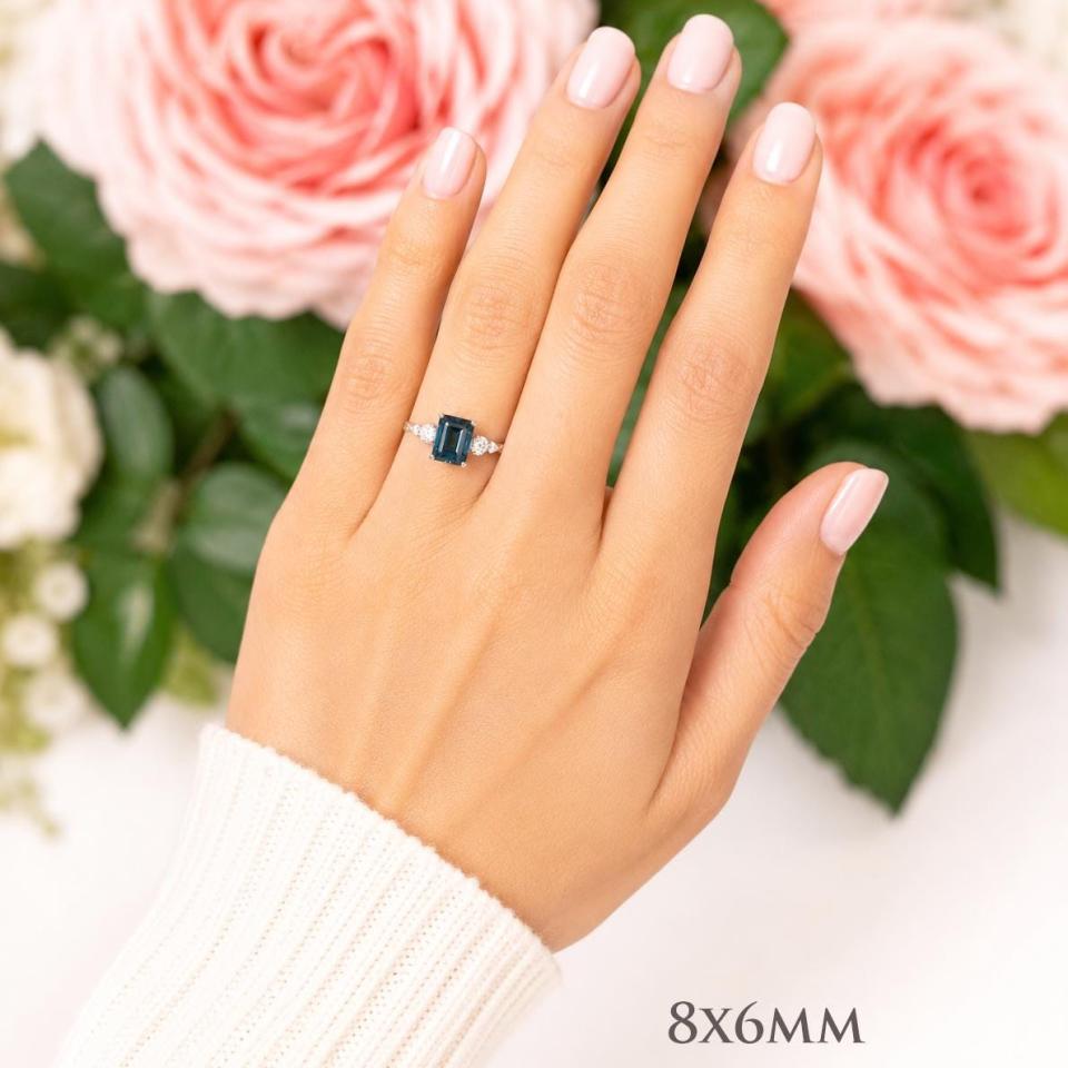A woman's hand wearing a sterling silver ring featuring an 8x6mm emerald cut London blue topaz flanked by a round moissanite on each side. Model image for size and styling reference — not a photo of the actual ring.