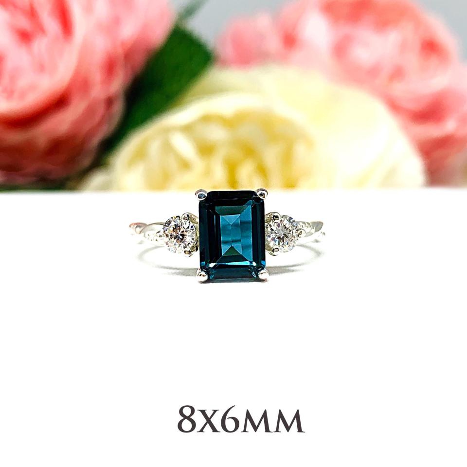 Close-up of an 8x6mm emerald-cut London Blue Topaz ring with moissanite side stones, set in a sterling silver twist band with milgrain detailing; soft floral background enhances the elegance.