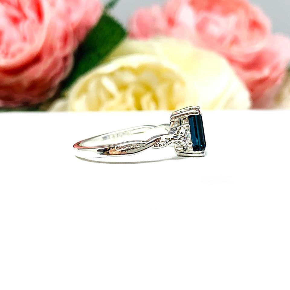 Close-up of an 8x6mm emerald-cut London Blue Topaz ring with moissanite side stones, set in a sterling silver twist band with milgrain detailing; soft floral background enhances the elegance.