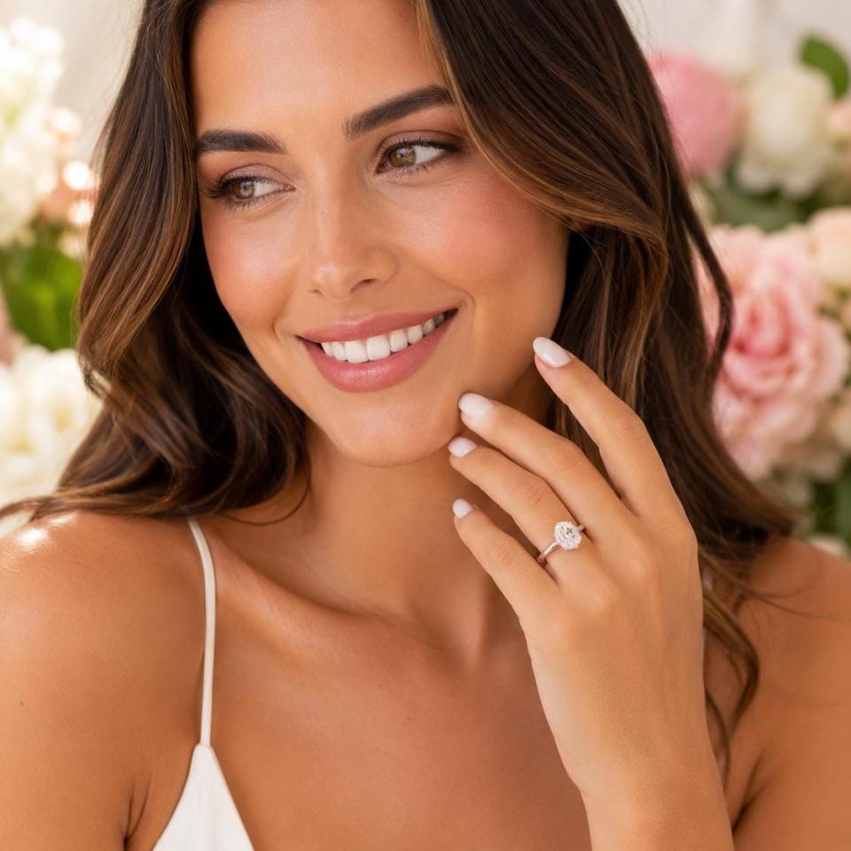 A woman wearing a sterling silver 6-prong solitaire engagement ring set with a 10x8mm near colorless oval cubic zirconia. Soft-focus pink and white flowers are in the background. Model image for size and styling reference — not a photo of the actual ring.