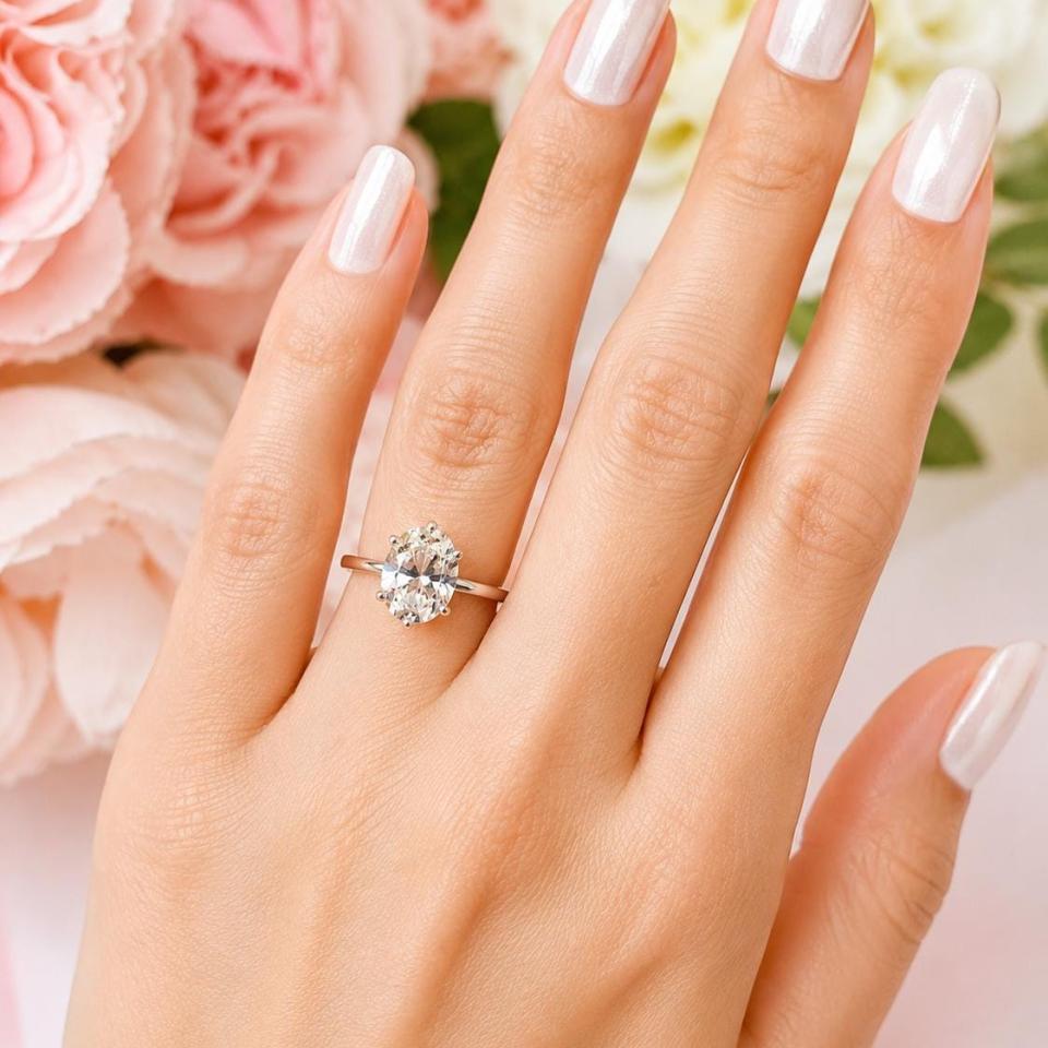 A woman's hand wearing a sterling silver 6-prong solitaire engagement ring set with a 10x8mm near colorless oval cubic zirconia. It is resting on a white surface with soft-focus pink and white flowers in the background. Model image for size and styling reference — not a photo of the actual ring.