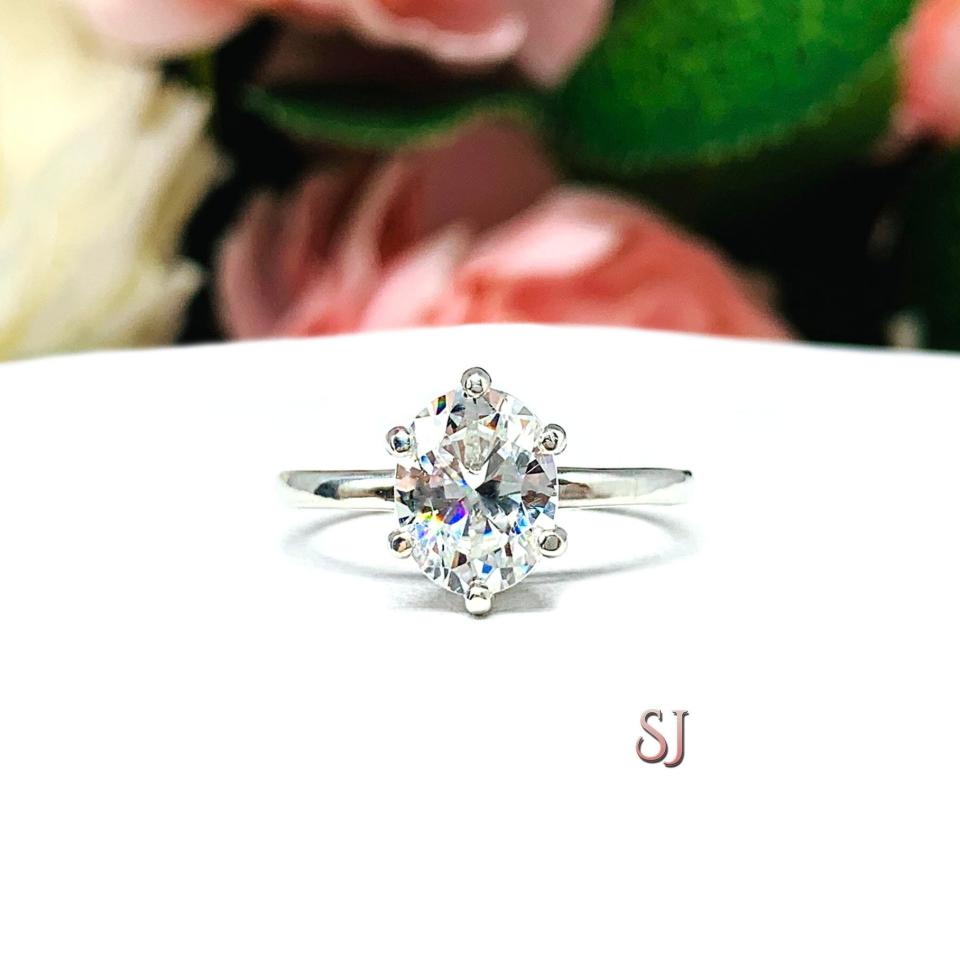 Sterling silver 6-prong solitaire engagement ring set with a 9x7mm near colorless oval cubic zirconia. It is resting on a white surface with soft-focus pink and white flowers in the background.