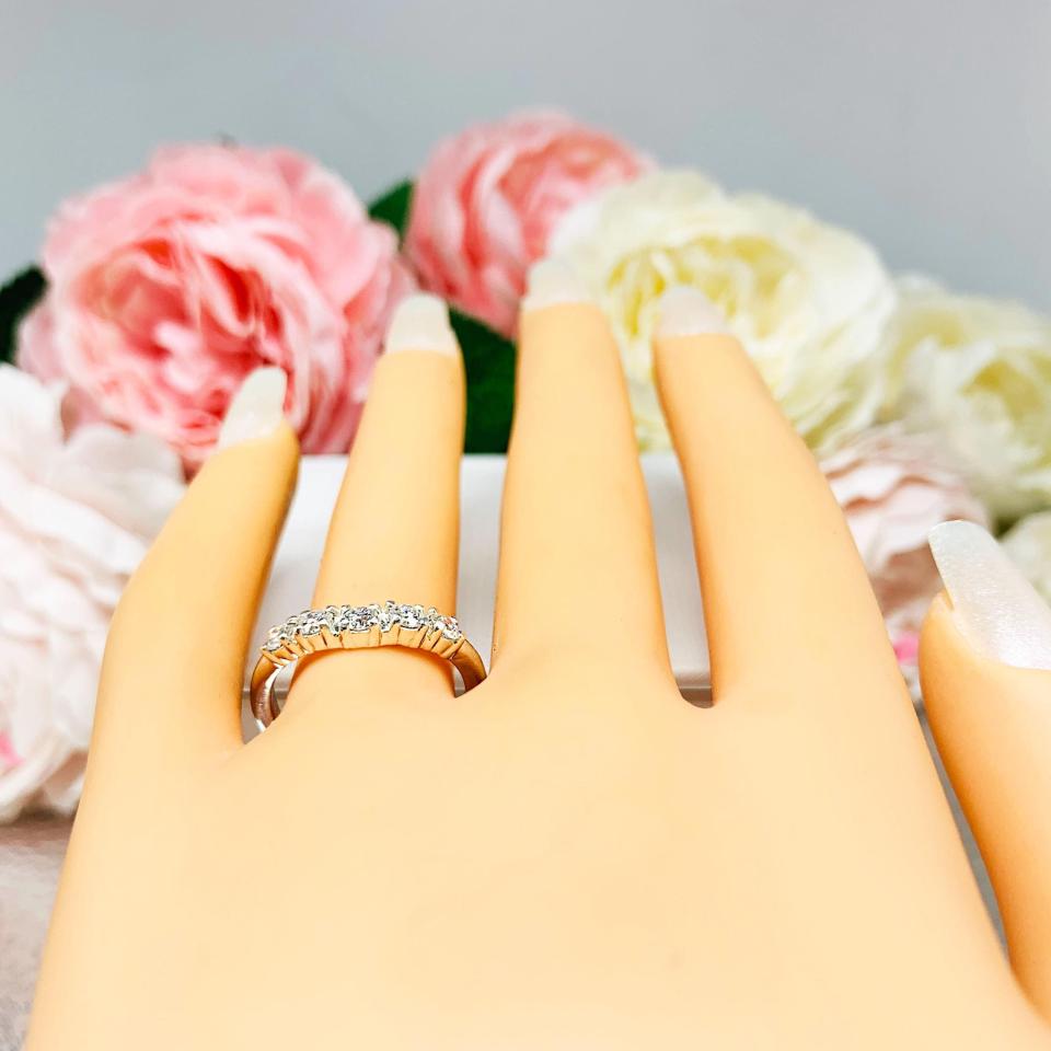 Mannequin hand with long white shimmery nails, displayed against a soft white-gray background with pink and white floral accents. The hand is wearing a sterling silver 5-stone curved wedding band featuring 3mm round precision faceted moissanite stones in prong settings.
