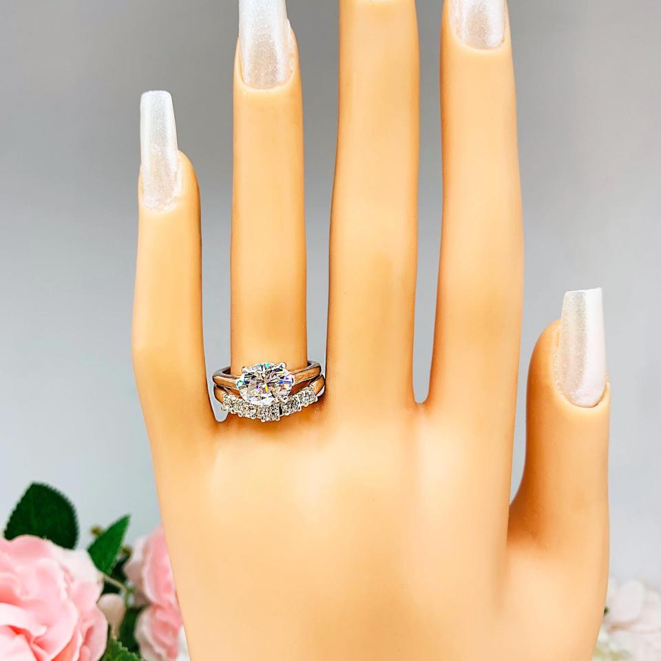 Mannequin hand with long white shimmery nails, displayed against a soft white-gray background with pink and white floral accents. The hand is wearing a sterling silver 5-stone curved wedding band featuring 3mm round precision faceted moissanite stones in prong settings. The ring is paired with an oval moissanite engagement ring. The oval ring is not included.