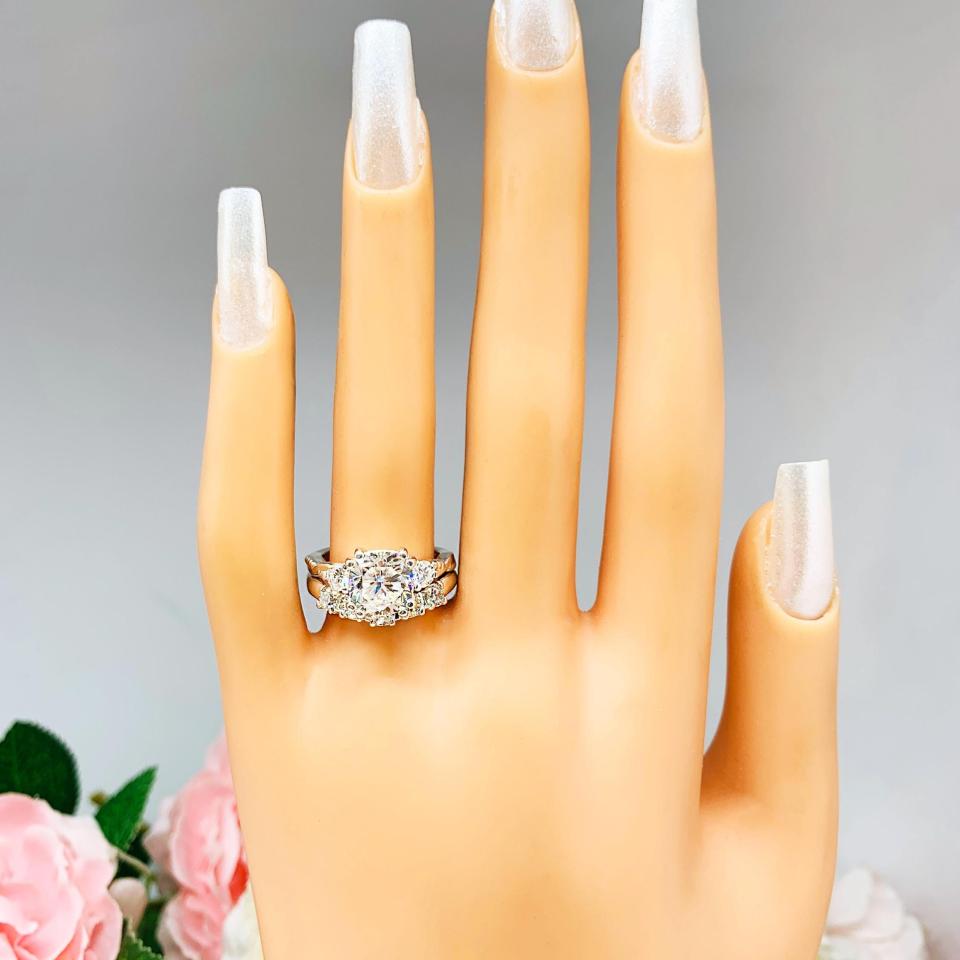 Mannequin hand with long white shimmery nails, displayed against a soft white-gray background with pink and white floral accents. The hand is wearing a sterling silver 5-stone curved wedding band featuring 3mm round precision faceted moissanite stones in prong settings. The ring is paired with a cushion with round side stone 3-stone moissanite engagement ring. This ring is not included.