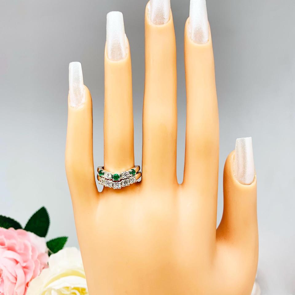 Mannequin hand with long white shimmery nails, displayed against a soft white-gray background with pink and white floral accents. The hand is wearing a sterling silver  five-stone curved wedding band with three 3mm round lab-created emeralds and two 3mm round DEF moissanites. Shown with a 5-stone curved moissanite band (not included).