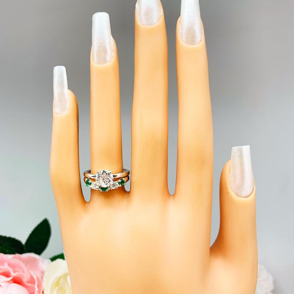 Mannequin hand with long white shimmery nails, displayed against a soft white-gray background with pink and white floral accents. The hand is wearing a sterling silver  five-stone curved wedding band with three 3mm round lab-created emeralds and two 3mm round DEF moissanites. Shown with a round moissanite engagement ring (not included).
