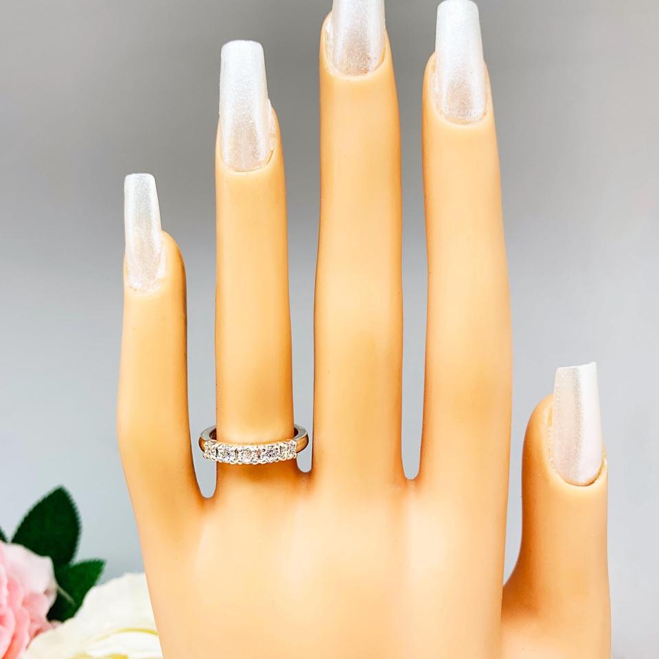Mannequin hand with long white shimmery nails, displayed against a soft white-gray background with pink and white floral accents. The hand is wearing a sterling silver stackable 5-stone wedding band featuring 3mm round authentic moissanite stones in prong settings.