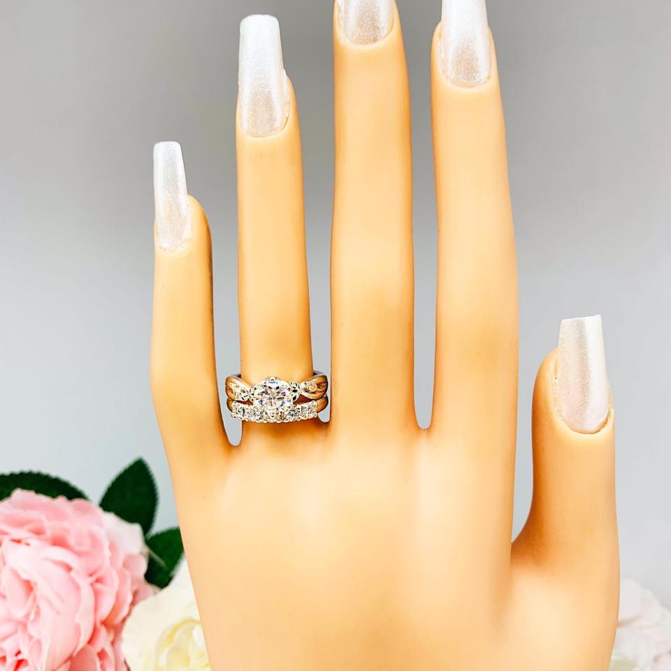 Mannequin hand with long white shimmery nails, displayed against a soft white-gray background with pink and white floral accents. The hand is wearing a sterling silver stackable 5-stone wedding band featuring 3mm round authentic moissanite stones in prong settings. The band is shown with a round moissanite engagement ring with leaf design. Engagement ring not included.