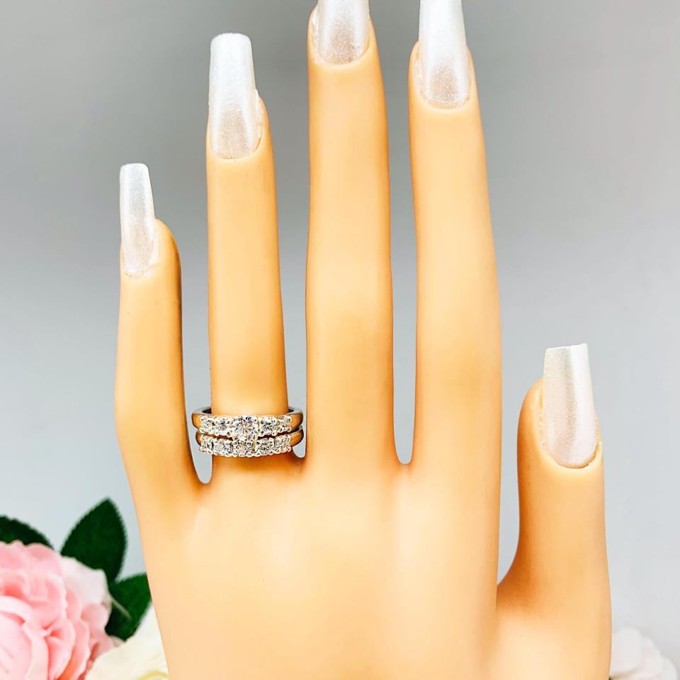 Mannequin hand with long white shimmery nails, displayed against a soft white-gray background with pink and white floral accents. The hand is wearing a sterling silver stackable 5-stone wedding band featuring 3mm round authentic moissanite stones in prong settings. The band is shown with a round moissanite engagement ring with graduated stone design. Engagement ring not included.