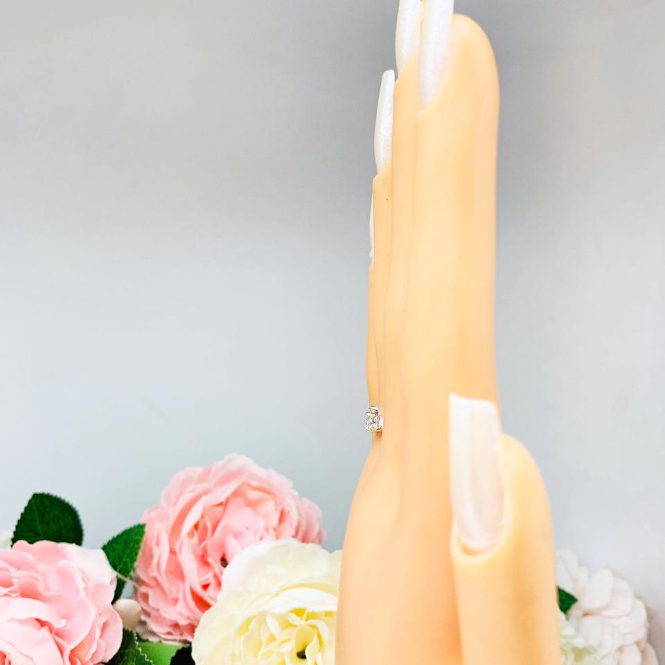 Mannequin hand with long white shimmery nails, displayed against a soft white-gray background with pink and white floral accents. The hand is wearing a sterling silver 5-stone curved wedding band featuring 3mm round precision faceted moissanite stones in prong settings.