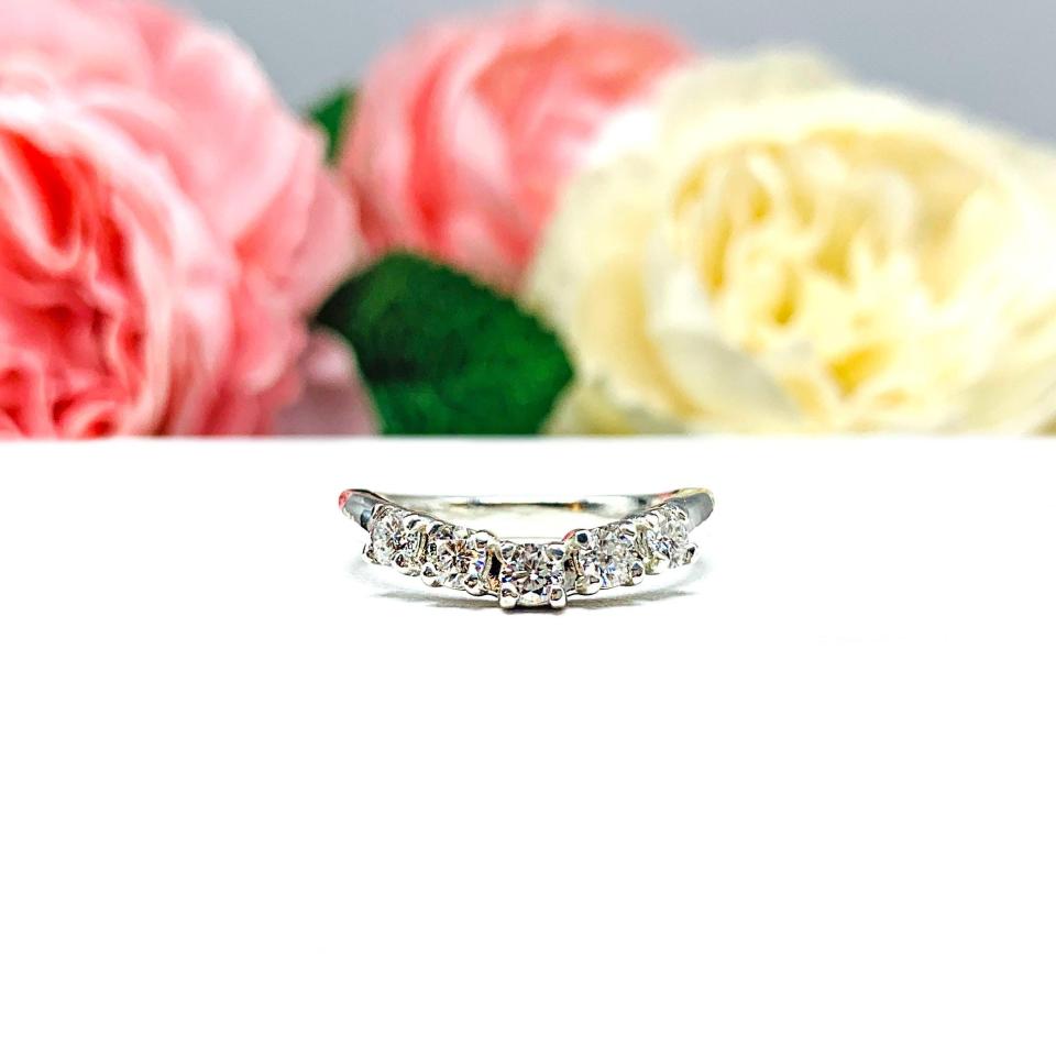Sterling silver 5-stone curved wedding band featuring 3mm round precision faceted moissanite stones in prong settings, displayed against a soft floral background.