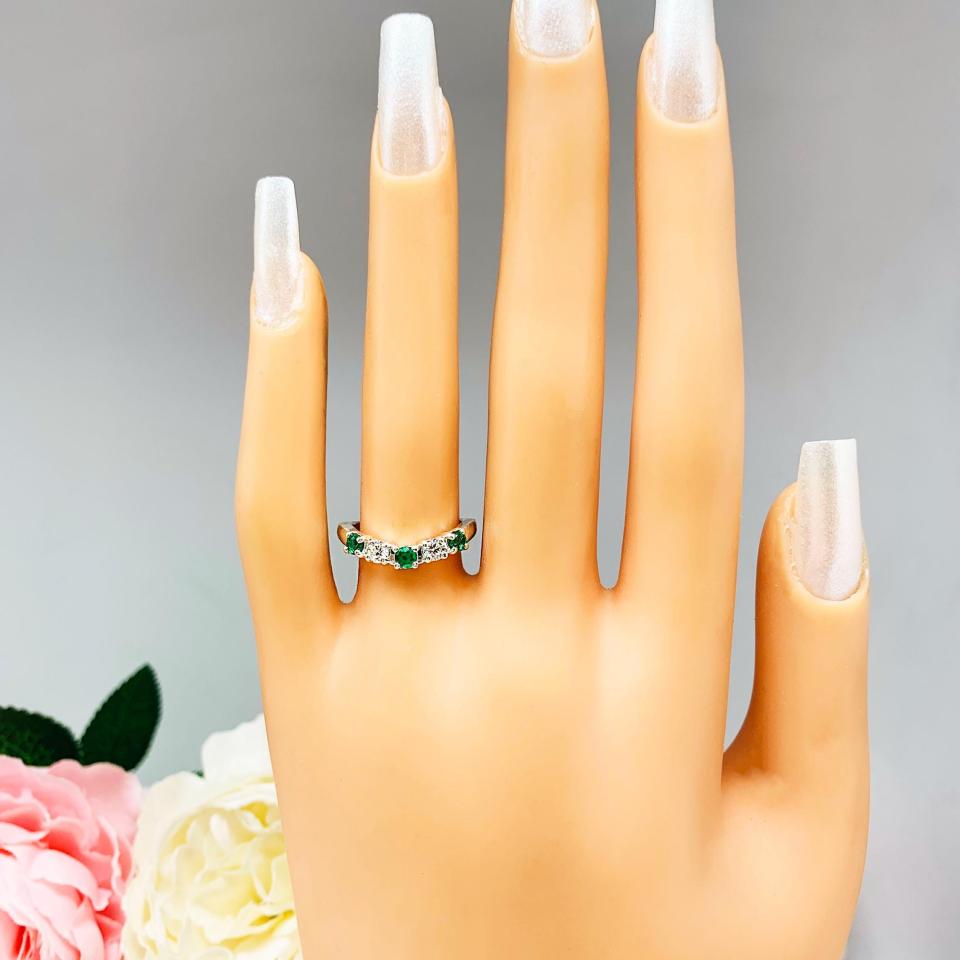 Mannequin hand with long white shimmery nails, displayed against a soft white-gray background with pink and white floral accents. The hand is wearing a sterling silver  five-stone curved wedding band with three 3mm round lab-created emeralds and two 3mm round DEF moissanites.