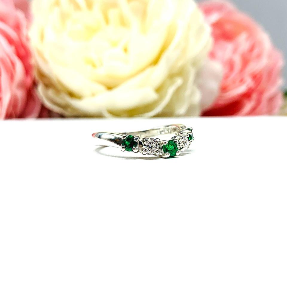 Sterling silver five-stone curved wedding band with three 3mm round lab-created emeralds and two 3mm round DEF moissanites, shown against a soft floral background.