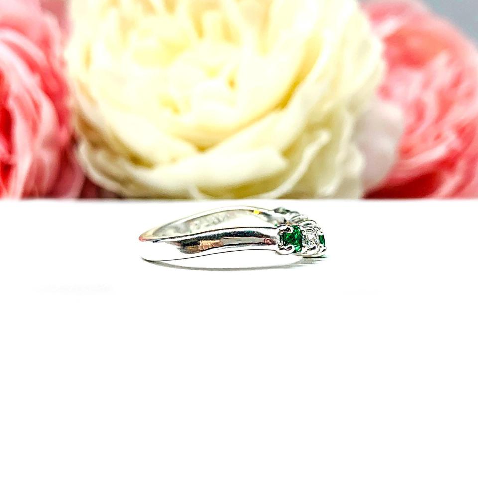 Sterling silver five-stone curved wedding band with three 3mm round lab-created emeralds and two 3mm round DEF moissanites, shown against a soft floral background.