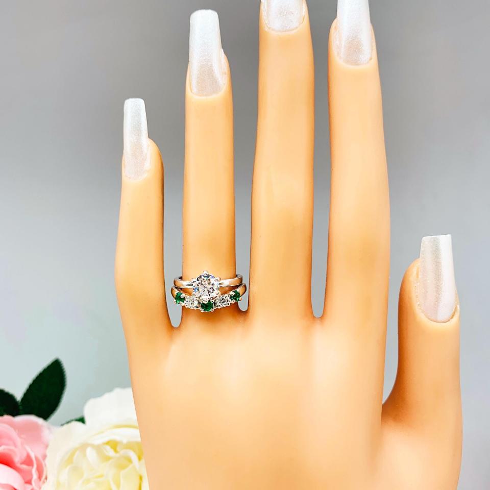 Mannequin hand with long white shimmery nails, displayed against a soft white-gray background with pink and white floral accents. The hand is wearing a sterling silver  five-stone curved wedding band with three 3mm round lab-created emeralds and two 3mm round DEF moissanites. Shown with a round moissanite engagement ring (not included).
