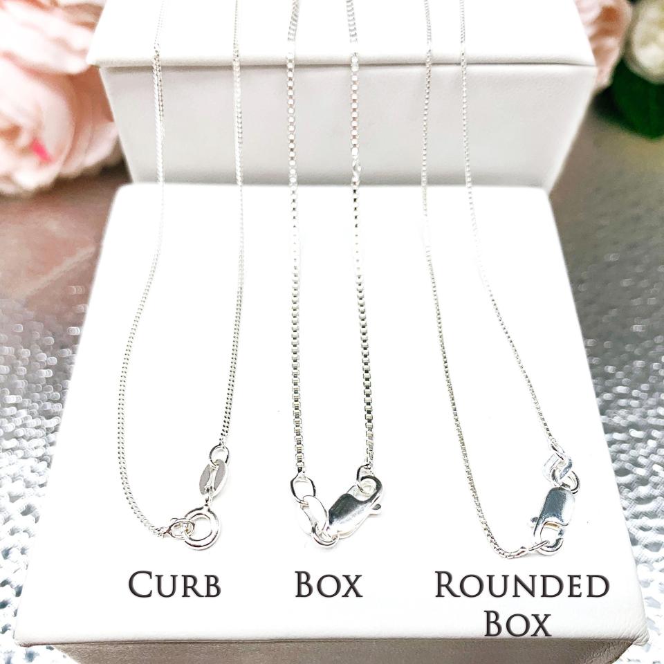 Three sterling silver chains displayed on a white jewelry stand, each labeled to show its distinct style: Curb, Box, and Rounded Box. The curb chain on the left has flat, interlocking links with a spring-ring clasp; the box chain in the center features square links and a lobster clasp; and the rounded box chain on the right has softer, curved links with a lobster clasp. Soft pink and cream flowers are blurred in the background, adding a delicate, elegant touch.
