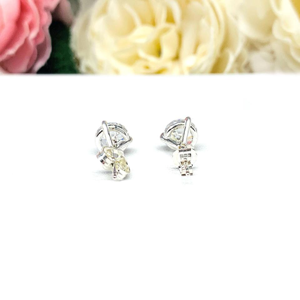 A pair of sterling silver 3-prong martini stud earrings with 6.5mm colorless old European cut cubic zirconias. The earrings are resting on a white surface with a background of pink and white flowers.