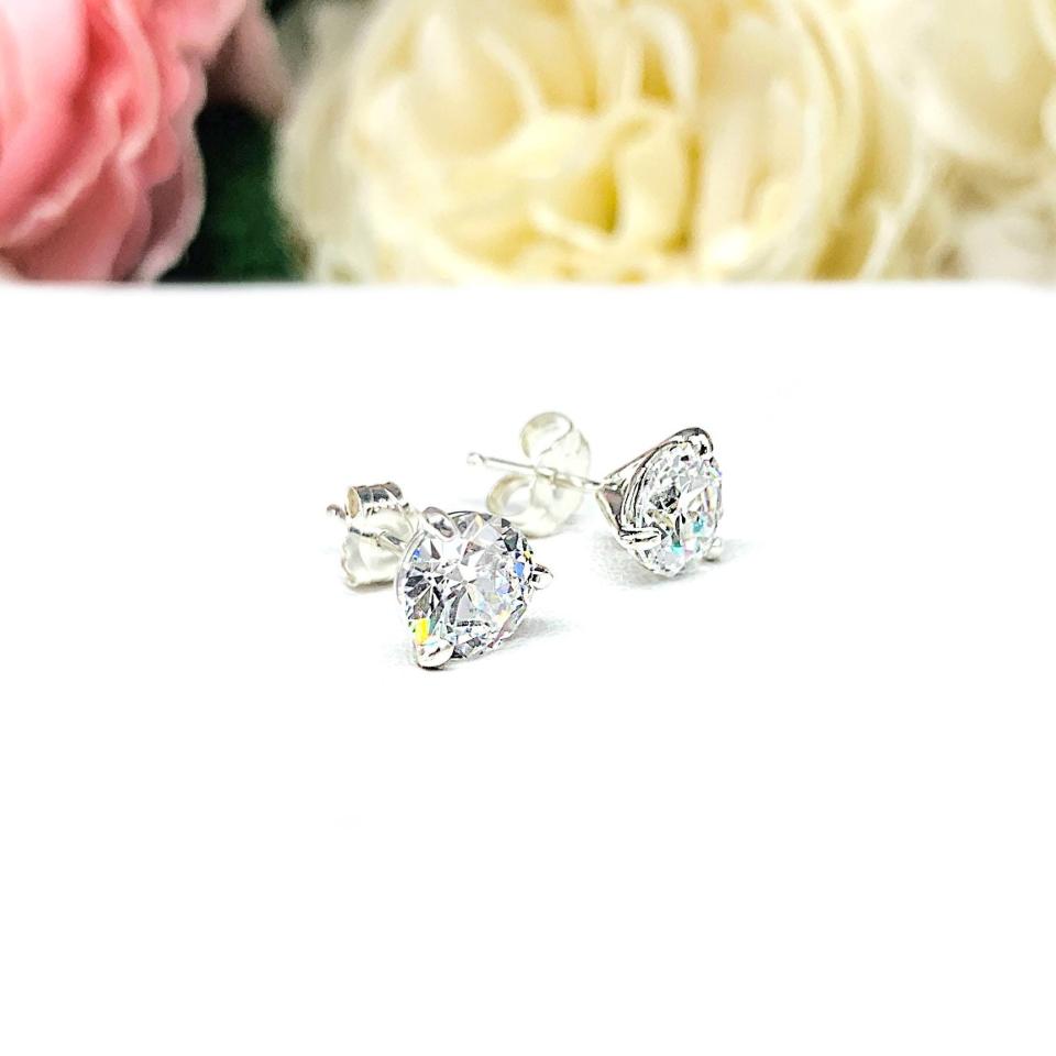 A pair of sterling silver 3-prong martini stud earrings with 6.5mm colorless old European cut cubic zirconias. The earrings are resting on a white surface with a background of pink and white flowers.