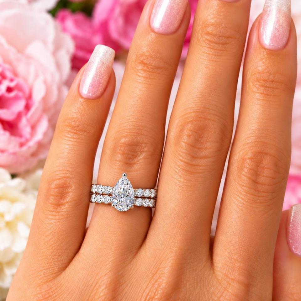 A woman's hand wearing an elegant sterling silver engagement ring set featuring a 10x7mm precision-cut pear cubic zirconia with pave CZ accents in a half eternity band. Shown with optional matching half-eternity band, designed to nest seamlessly. Model image for size and styling reference — not a photo of the actual ring.
