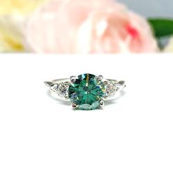 Sterling silver three-stone ring featuring a vivid green moissanite center stone flanked by two smaller white moissanites. The band has a delicate milgrain twist design and a polished finish. Displayed against a soft-focus floral background, the ring sparkles with contrast and elegance.