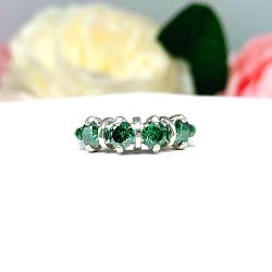 Sterling silver ring with four round emerald green moissanite stones in a row, displayed against a soft-focus background of pink and white flowers. The moissanites are vivid and dazzling, and the band has a smooth, polished finish.