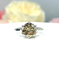 Sterling silver engagement ring with a round light champagne moissanite in a six-prong trellis setting. The non-plated silver band is 2.5mm wide and polished to a soft shine. Displayed against a soft-focus floral background, the moissanite glows with warm, golden undertones.