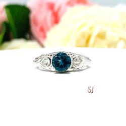 A sterling silver ring with a richly saturated 6mm round London blue topaz gemstone set in a delicate filigree design. Displayed on a white surface with a pastel floral background. The letters 'SJ' appear in the bottom right corner.