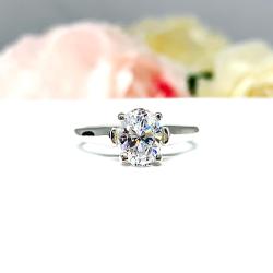 Sterling silver solitaire cathedral engagement ring featuring a dazzling 8x6mm brilliant cut oval cubic zirconia. The ring is elegantly displayed on a white surface with a background of soft-focus pink and white flowers.