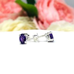 A pair of four prong dainty silver oval stud earrings with purple African amethyst gemstones. The basket earrings are on a white surface with the left earring facing forward to showcase the crisp clarity and deep purple color of the gemstones and the right earring facing toward the right to display the post at the top and the butterfly friction clasp.