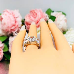 Mannequin hand with long white shimmery nails, displayed against a soft white-gray background with pink and white floral accents. The hand is wearing a sterling silver 3-stone engagement ring featuring an 8x6mm-10x8mm radiant cut cubic zirconia center stone with two smaller radiant CZ side stones. The highly polished wide band tapers for comfort.