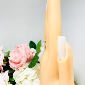 Mannequin hand with long white shimmery nails, displayed against a soft white-gray background with pink and white floral accents. The hand is wearing a sterling silver 3-stone engagement ring featuring an 8x6mm-10x8mm radiant cut cubic zirconia center stone with two smaller radiant CZ side stones. The highly polished wide band tapers for comfort.