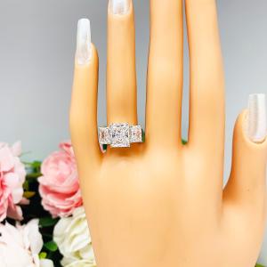 Mannequin hand with long white shimmery nails, displayed against a soft white-gray background with pink and white floral accents. The hand is wearing a sterling silver 3-stone engagement ring featuring an 8x6mm-10x8mm radiant cut cubic zirconia center stone with two smaller radiant CZ side stones. The highly polished wide band tapers for comfort.