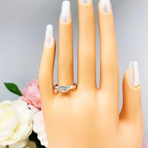 Mannequin hand with long white shimmery nails, displayed against a soft white-gray background with pink and white floral accents. The hand is wearing a 6-prong sterling silver, horizontal east west set engagement ring featuring a precision-faceted near colorless (G/H color) marquise cubic zirconia.