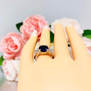 Mannequin hand with long white shimmery nails, displayed against a soft white-gray background with pink and white floral accents. The hand is wearing a sterling silver 3-stone ring with a round lab-created blue sapphire center flanked by smaller round brilliant cut cubic zirconias on each side.