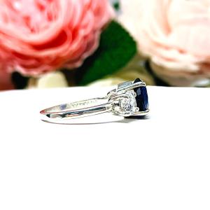 Sterling silver 3-stone ring with a round lab-created blue sapphire center flanked by smaller round brilliant cut cubic zirconias on each side. The ring is set against a white surface with a background of pink and white flowers.