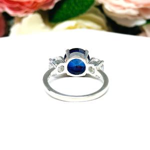 Sterling silver 3-stone ring with a round lab-created blue sapphire center flanked by smaller round brilliant cut cubic zirconias on each side. The ring is set against a white surface with a background of pink and white flowers.