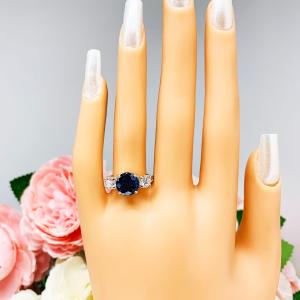 Mannequin hand with long white shimmery nails, displayed against a soft white-gray background with pink and white floral accents. The hand is wearing a sterling silver 3-stone ring with a round lab-created blue sapphire center flanked by smaller round brilliant cut cubic zirconias on each side.