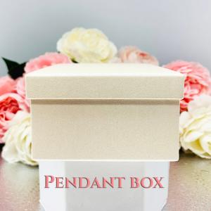An ivory color square, cardboard box with a textured surface. The box is sitting on a white surface with the words 'PENDANT BOX' printed in pink letters.