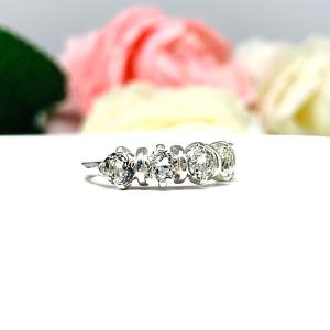 A sterling silver ring featuring five 4mm round white topaz gemstones in a row. The ring has rounded bars separating each stone. Displayed on a white surface with pastel floral background.