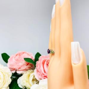 Mannequin hand with long white shimmery nails, displayed against a soft white-gray background with pink and white floral accents. The hand is wearing a sterling silver ring featuring a vibrant 8mm round African amethyst flanked by two pear-cut peridot side stones. The rich purple and bright green gemstones create a striking contrast, set against a soft floral background that enhances the ring’s natural elegance.