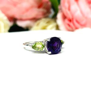Sterling silver ring featuring a vibrant 8mm round African amethyst flanked by two pear-cut peridot side stones. The rich purple and bright green gemstones create a striking contrast, set against a soft floral background that enhances the ring’s natural elegance.